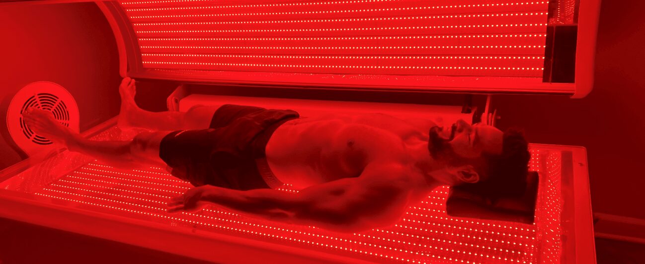 Man lying in a red-lit tanning bed wearing shorts.