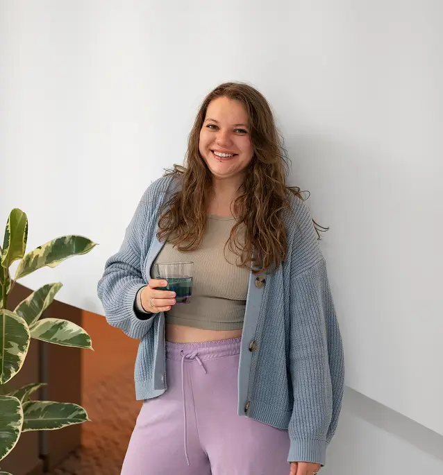 A smiling woman in casual attire stands beside a plant against a white wall.